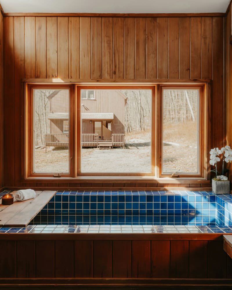 A timber bathing room with a tiled bath beneath a wide window, a folded towel and a single orchid.