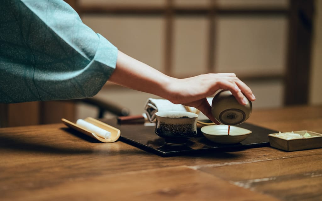 A host in a teal kimono sleeve pours tea at a low timber table.