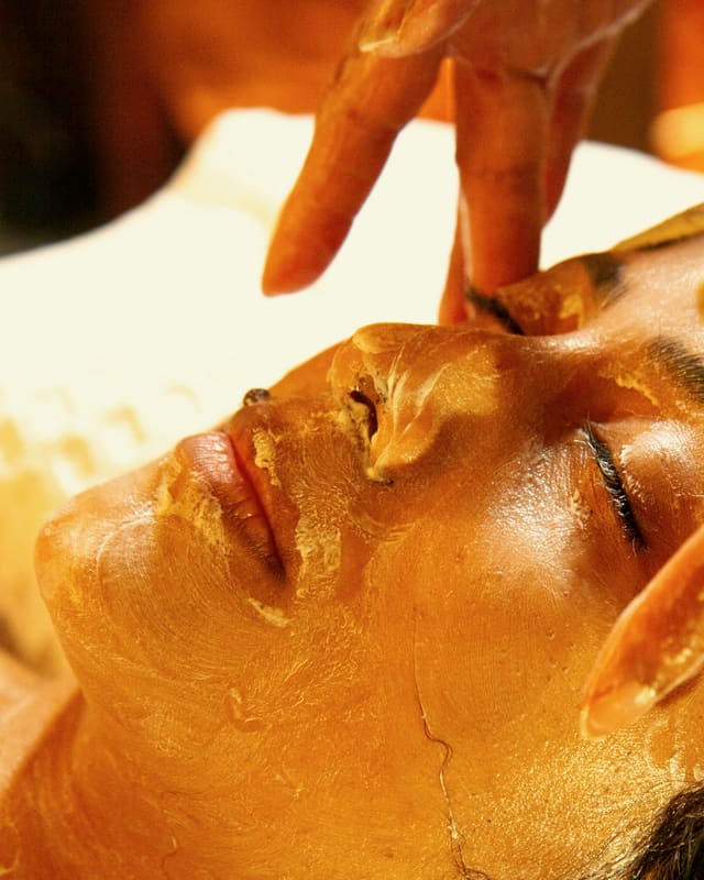 A therapist’s hands smooth a golden mask across a guest’s face, eyes closed.