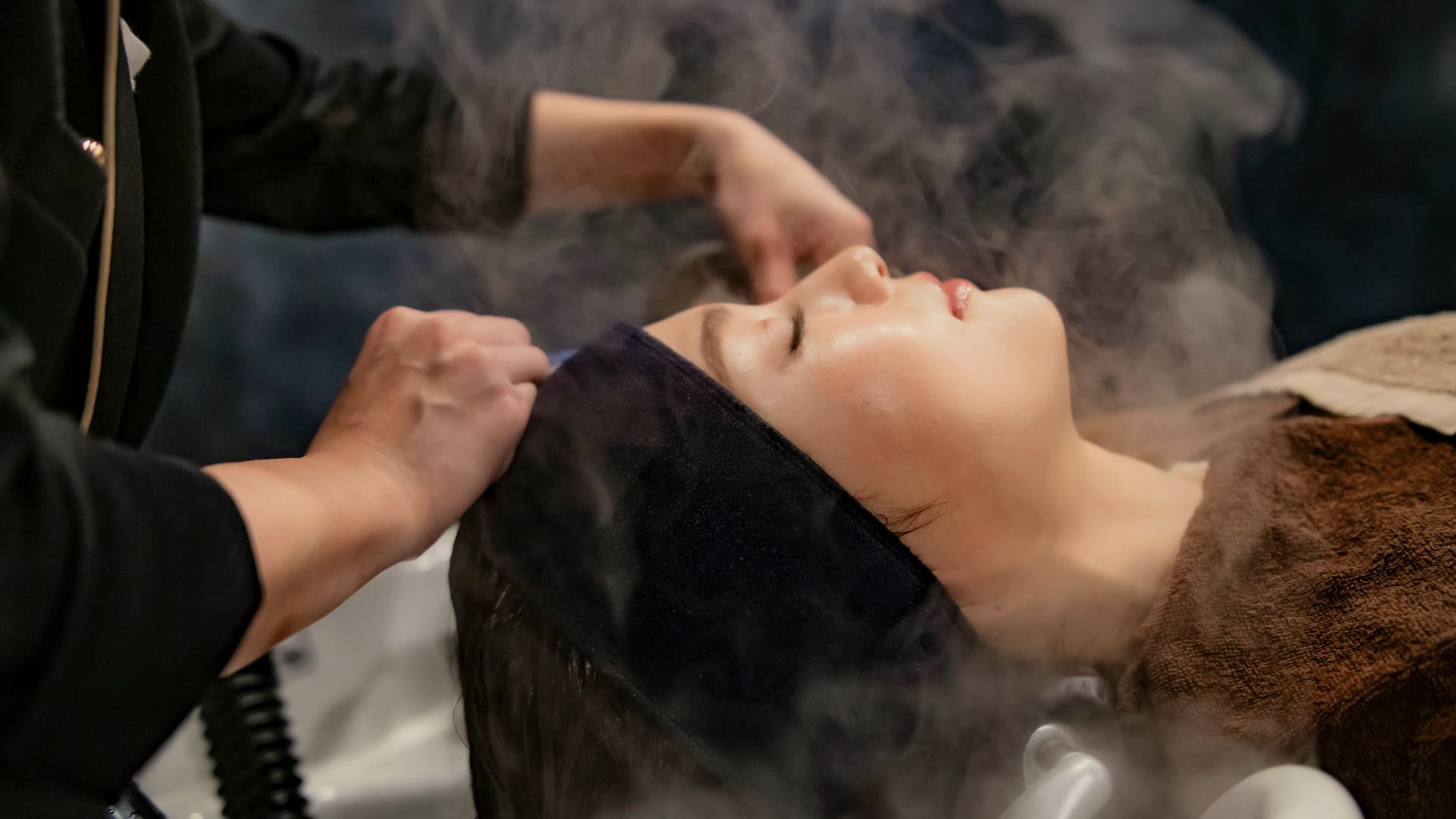 A therapist rinses a guest’s hair over a basin as steam rises in a dim treatment room.