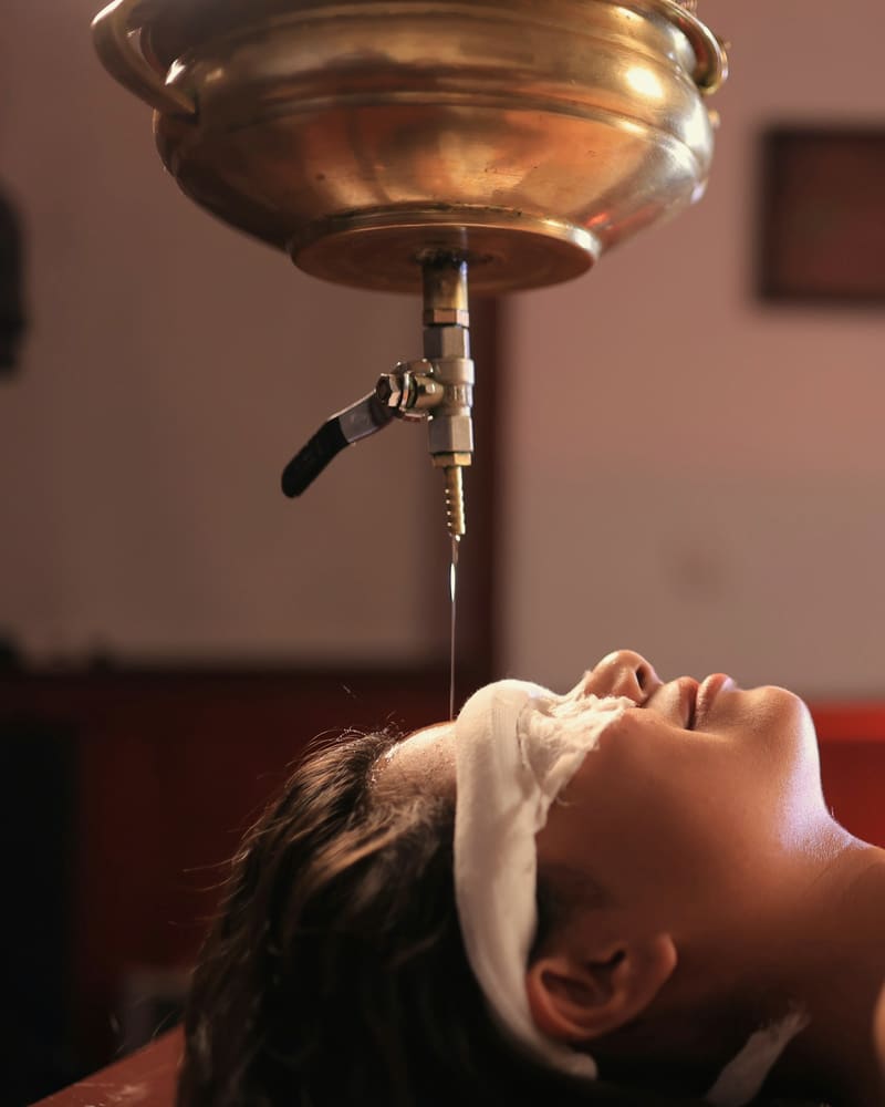 A guest lies still, eyes covered, beneath a copper vessel as warm oil falls toward her forehead.